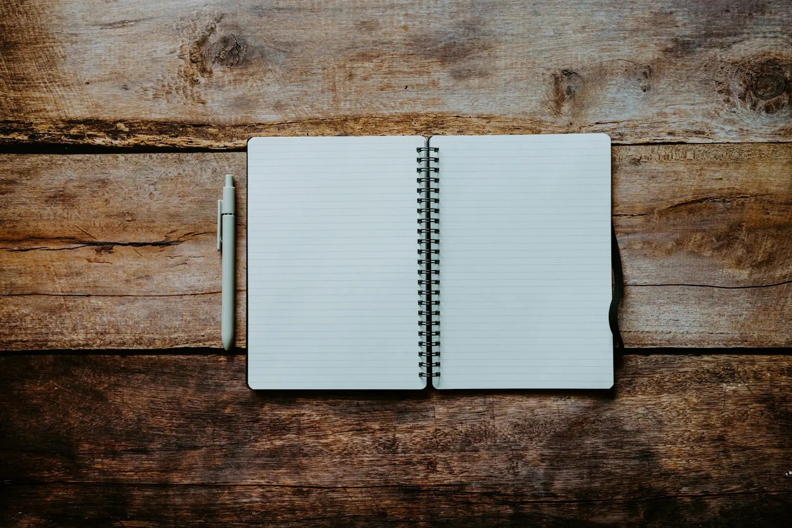 An open spiral-bound notebook with blank lined pages and a pen resting beside it on a wooden table