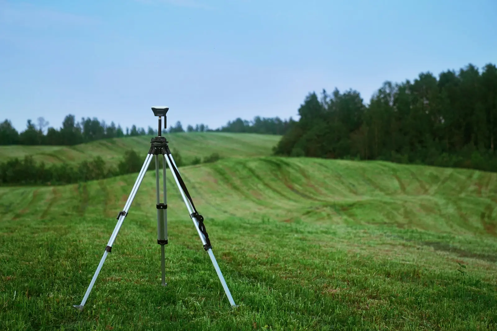 A tripod-mounted GPS surveying instrument standing in an open green field with rolling hills behind it