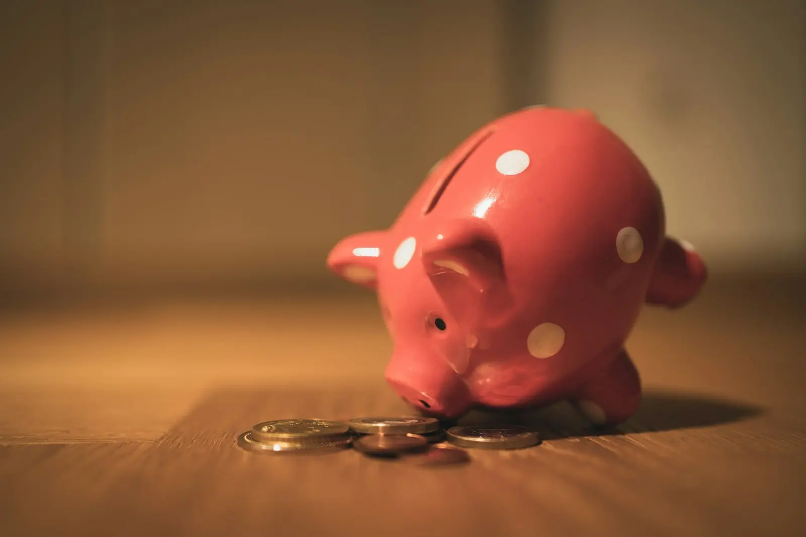 A pink polka-dot piggy bank standing beside a small pile of coins on a wooden floor