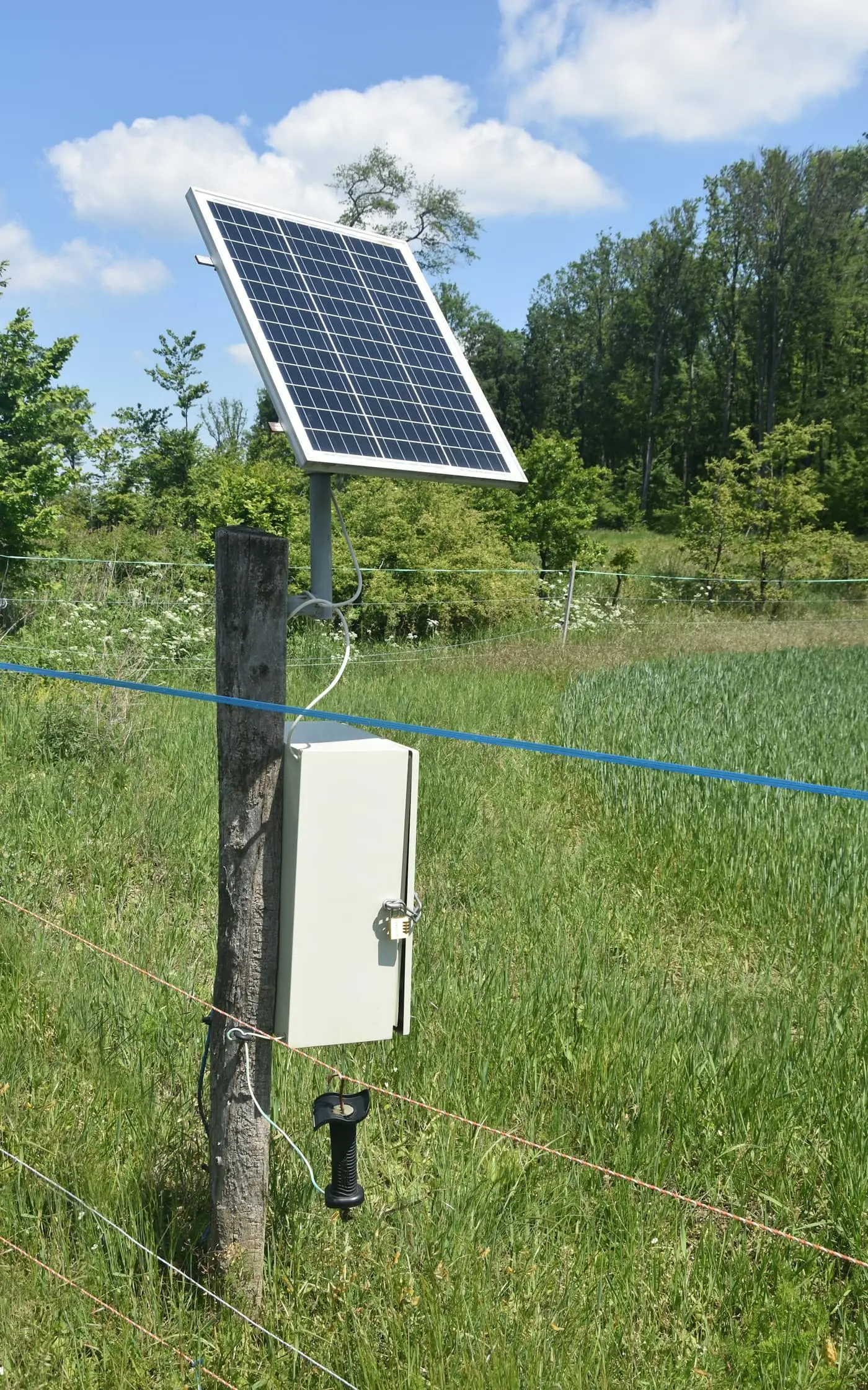 A solar panel on a pole beside a locked grey control box next to a wire fence on green farmland, with trees behind