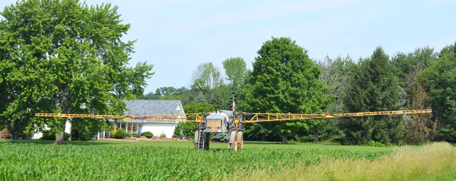 A self-propelled boom sprayer with wide extended arms parked in a field near a farmhouse
