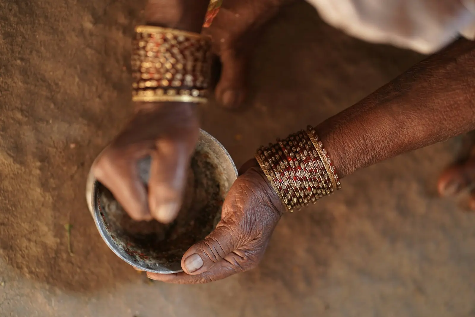 Bangled hands mixing a dark paste in a metal bowl, outdoors on bare earth