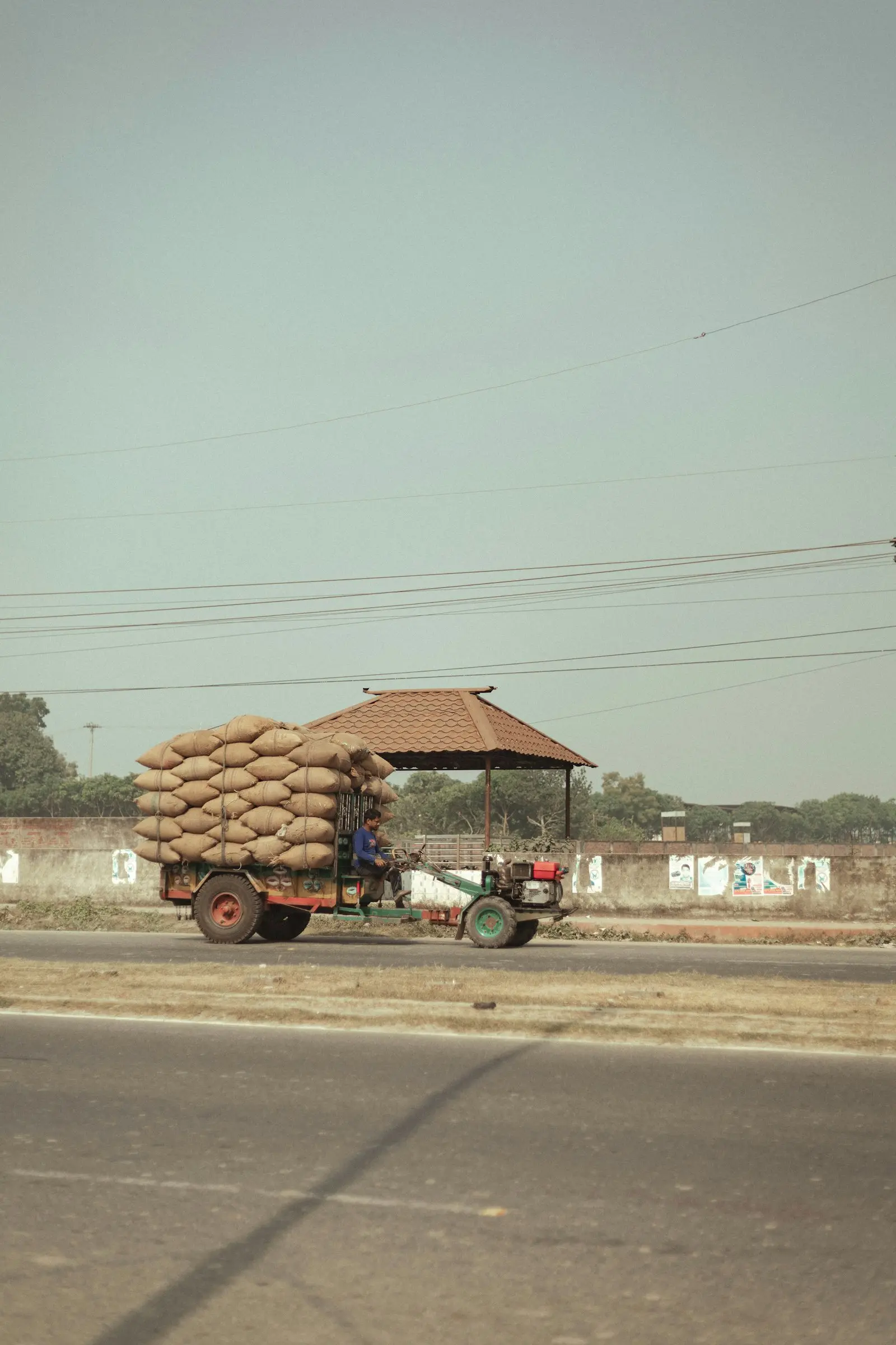 A small motorised cart heavily loaded with jute sacks of grain, driven along a rural road