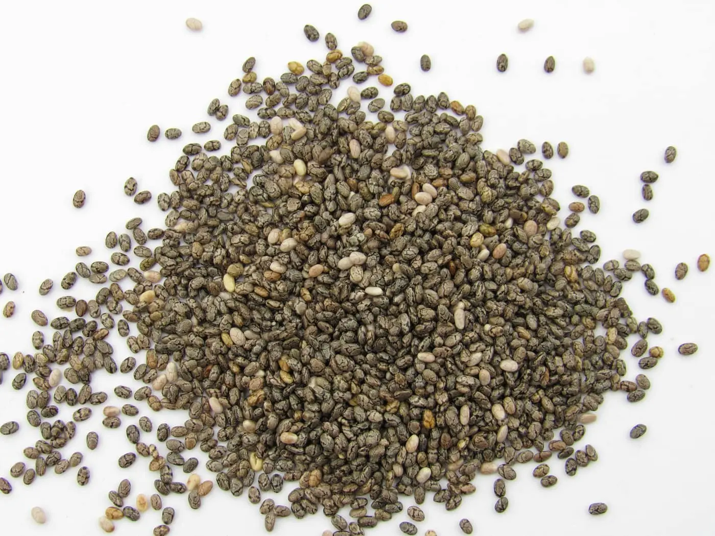 A close-up pile of tiny dark, mottled chia seeds heaped on a plain white surface