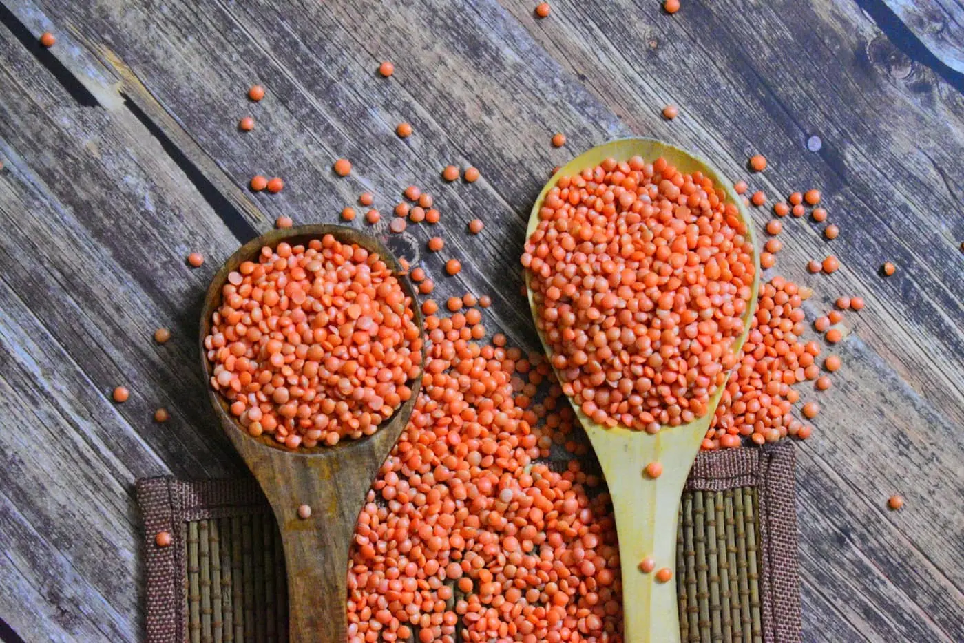 Two wooden spoons piled high with red lentils, spilling across a weathered wooden table