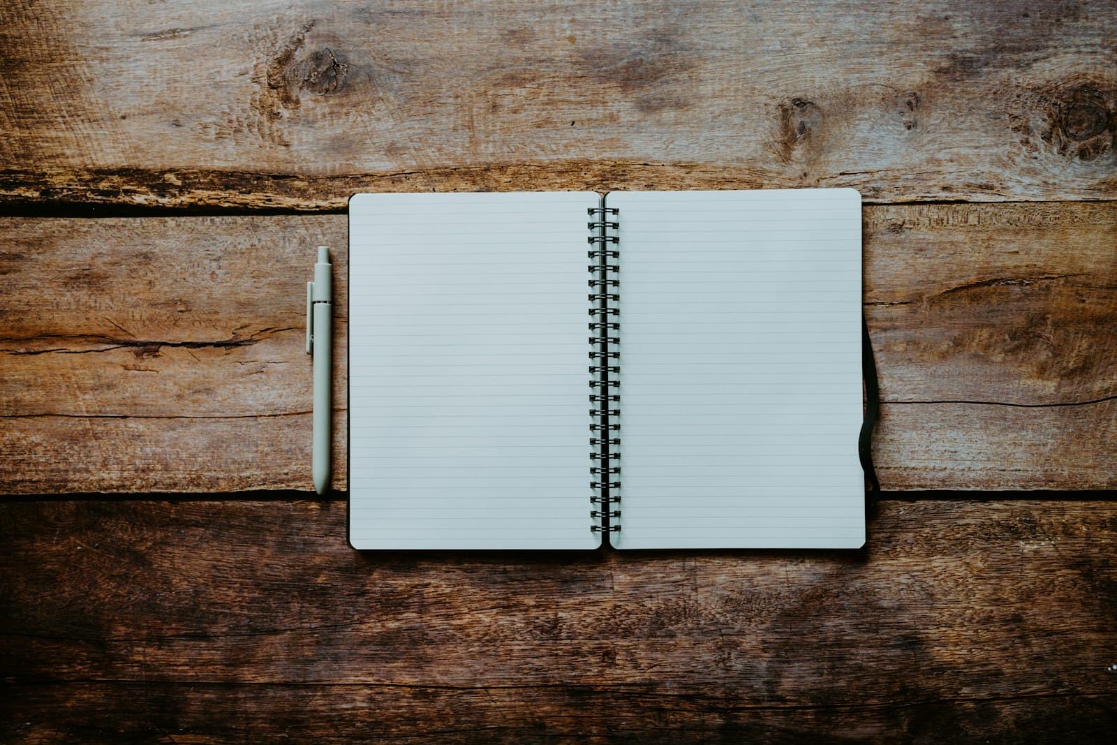 An open spiral-bound notebook with blank lined pages and a pen resting beside it on a wooden table