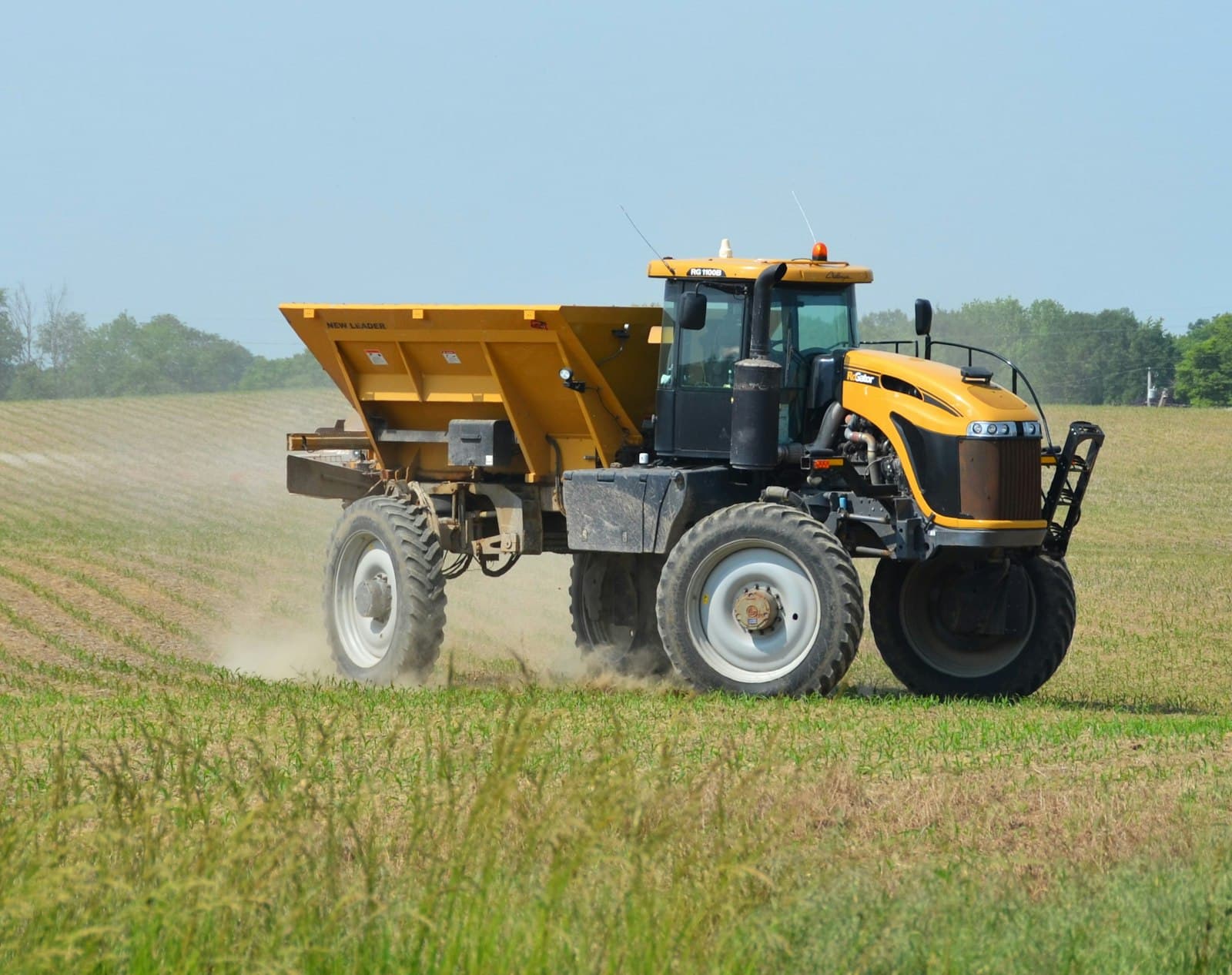 A yellow tractor towing a granular fertiliser spreader across a field, kicking up dust