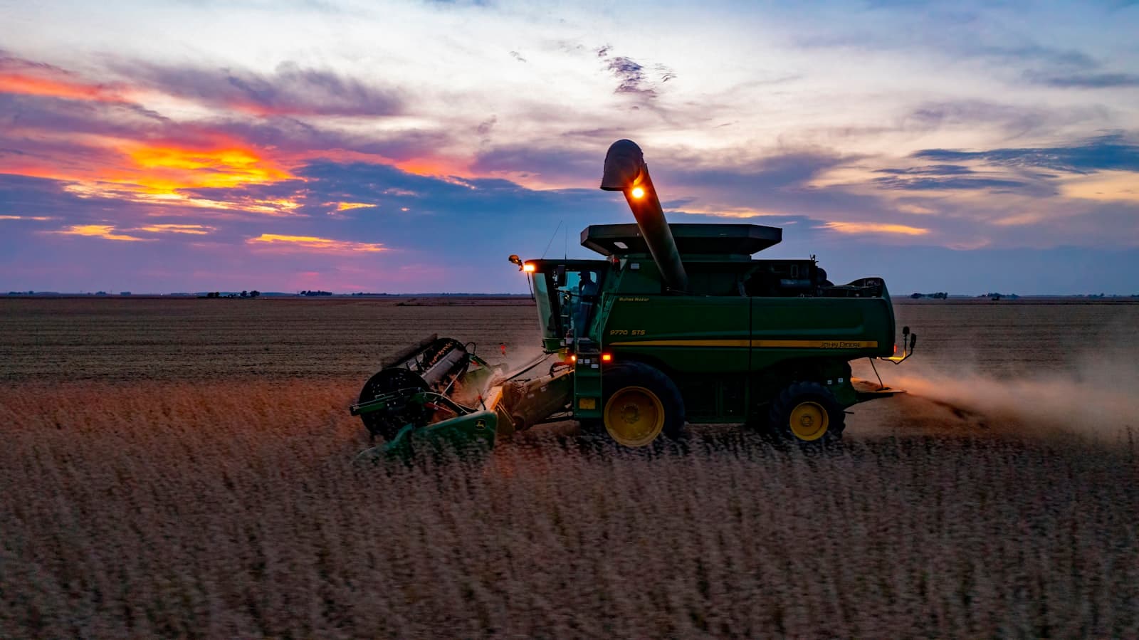 A green combine harvester working a golden field at sunset with dust rising behind it