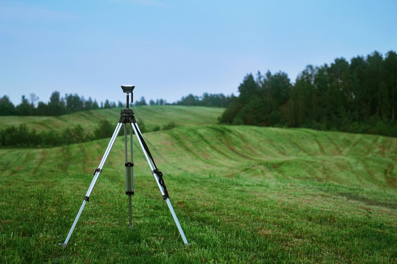 A tripod-mounted GPS surveying instrument standing in an open green field with rolling hills behind it