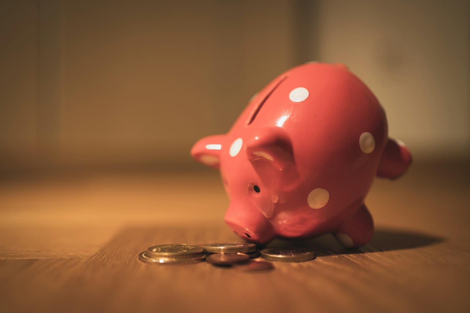 A pink polka-dot piggy bank standing beside a small pile of coins on a wooden floor