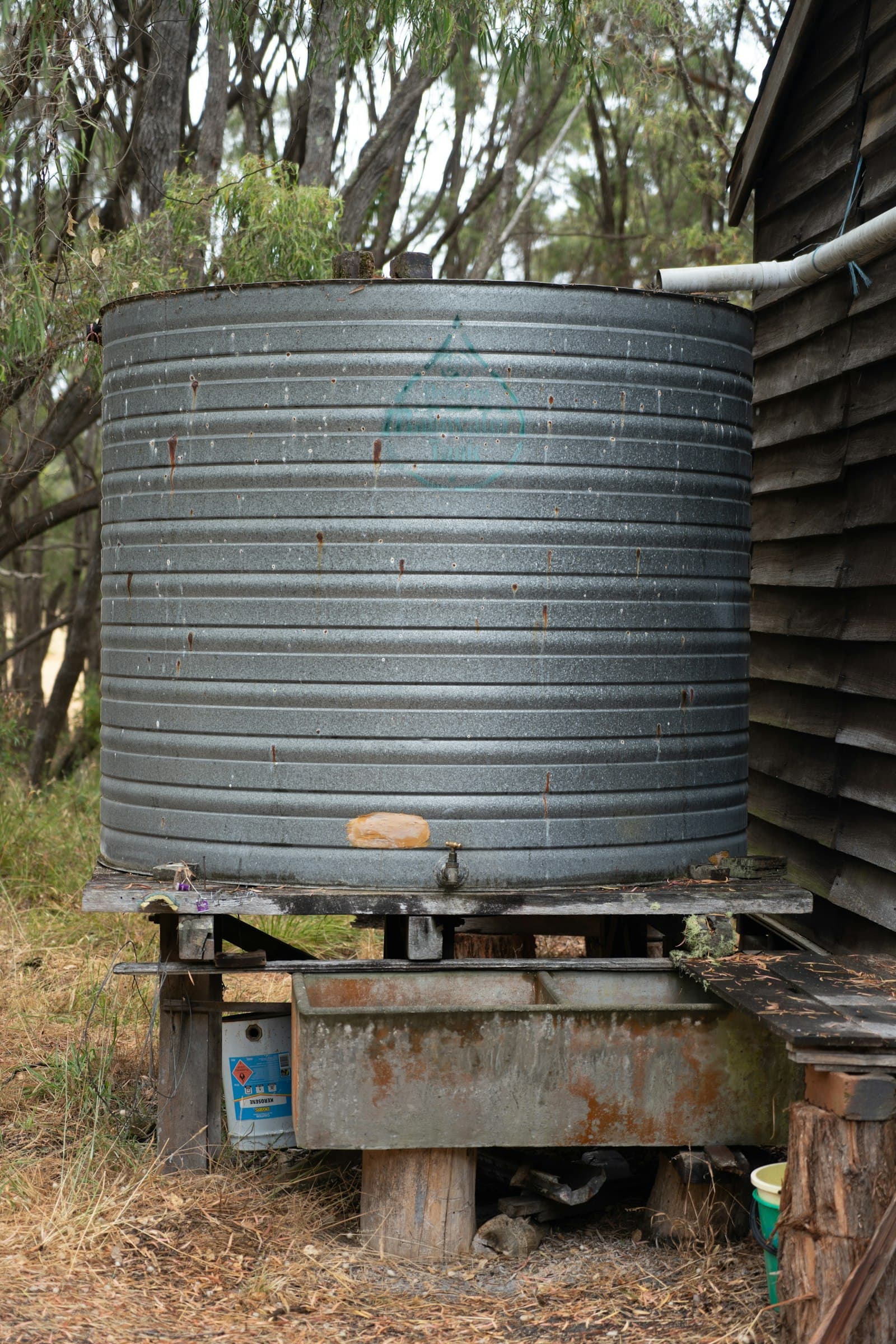 A large corrugated metal rainwater storage tank standing on a wooden platform beside a shed