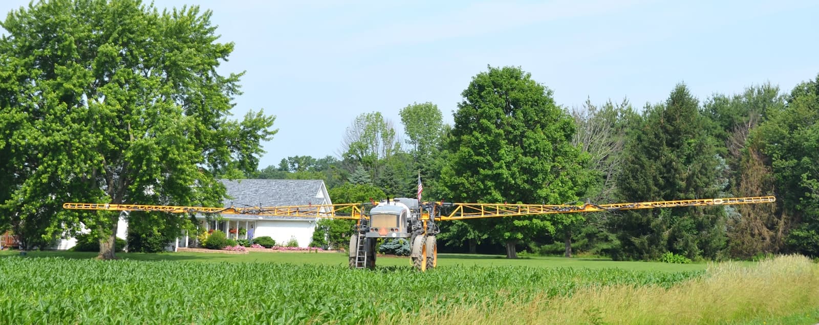 A self-propelled boom sprayer with wide extended arms parked in a field near a farmhouse
