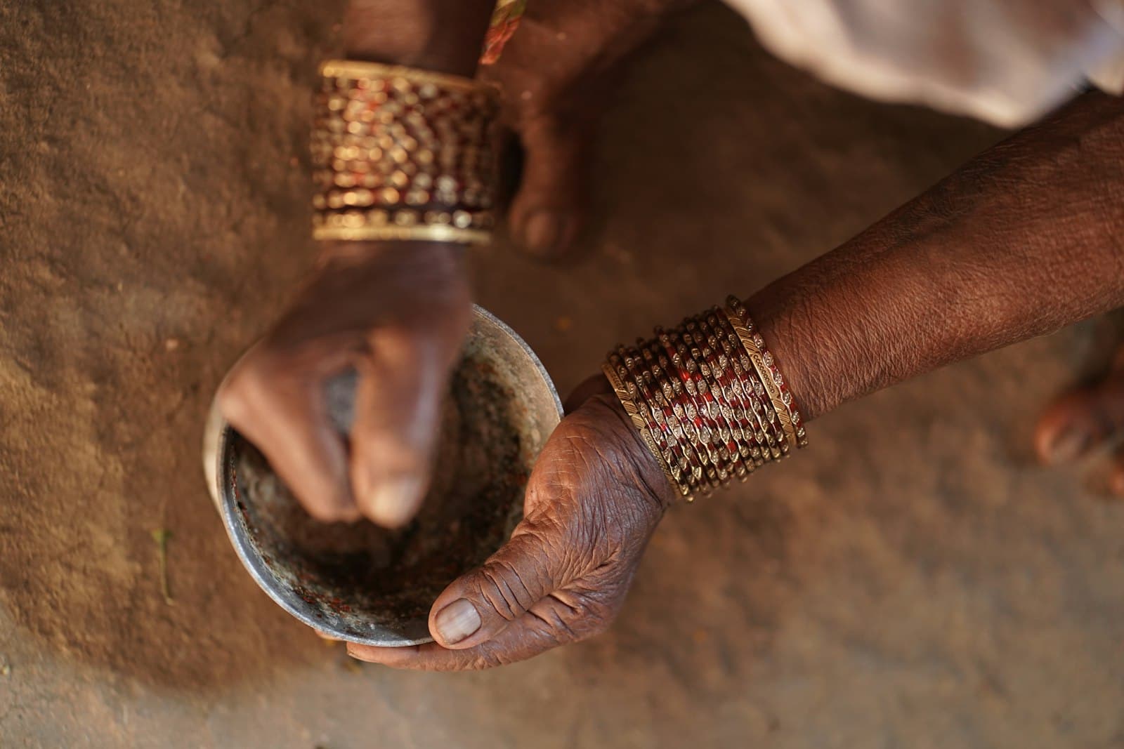 Bangled hands mixing a dark paste in a metal bowl, outdoors on bare earth