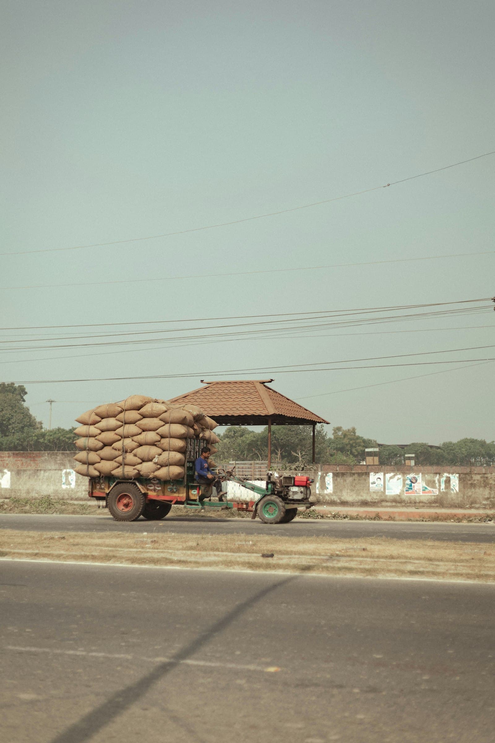 A small motorised cart heavily loaded with jute sacks of grain, driven along a rural road