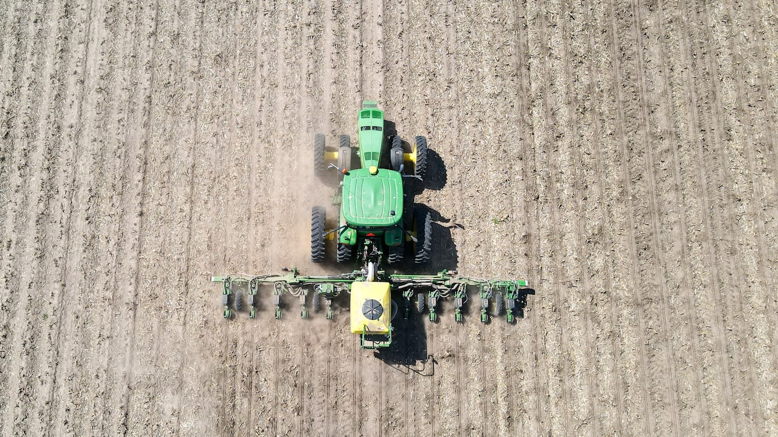 Aerial view of a tractor towing a wide seed drill directly across a residue-covered field, raising a trail of dust