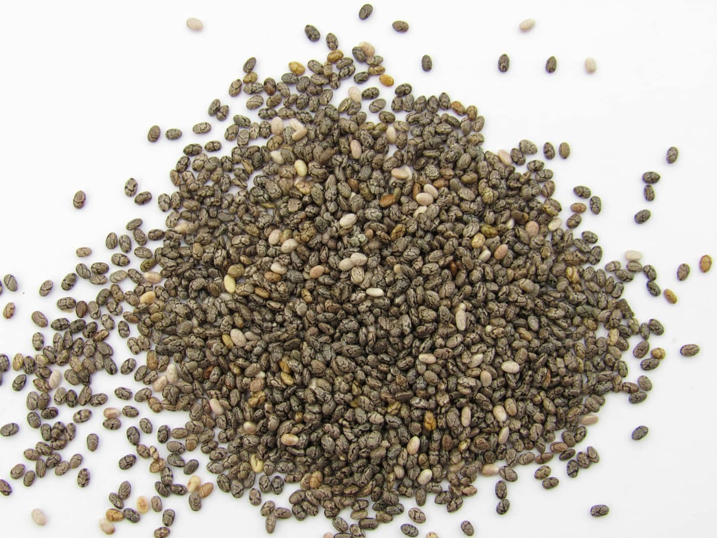 A close-up pile of tiny dark, mottled chia seeds heaped on a plain white surface