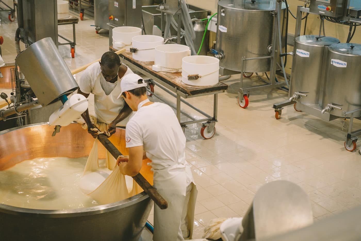 Two workers in white uniforms stirring curd in a large copper vat inside a dairy processing plant