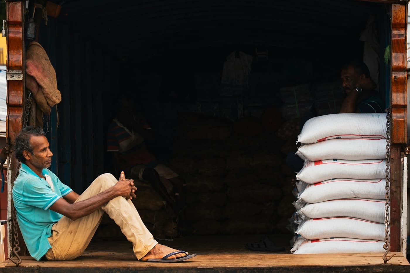A man sitting at the back of a loading dock beside a tall stack of sealed white grain sacks