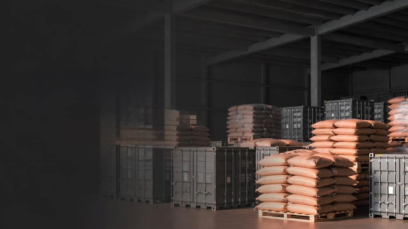 Tall stacks of grain sacks on wooden pallets inside a large warehouse, with shipping containers in the background