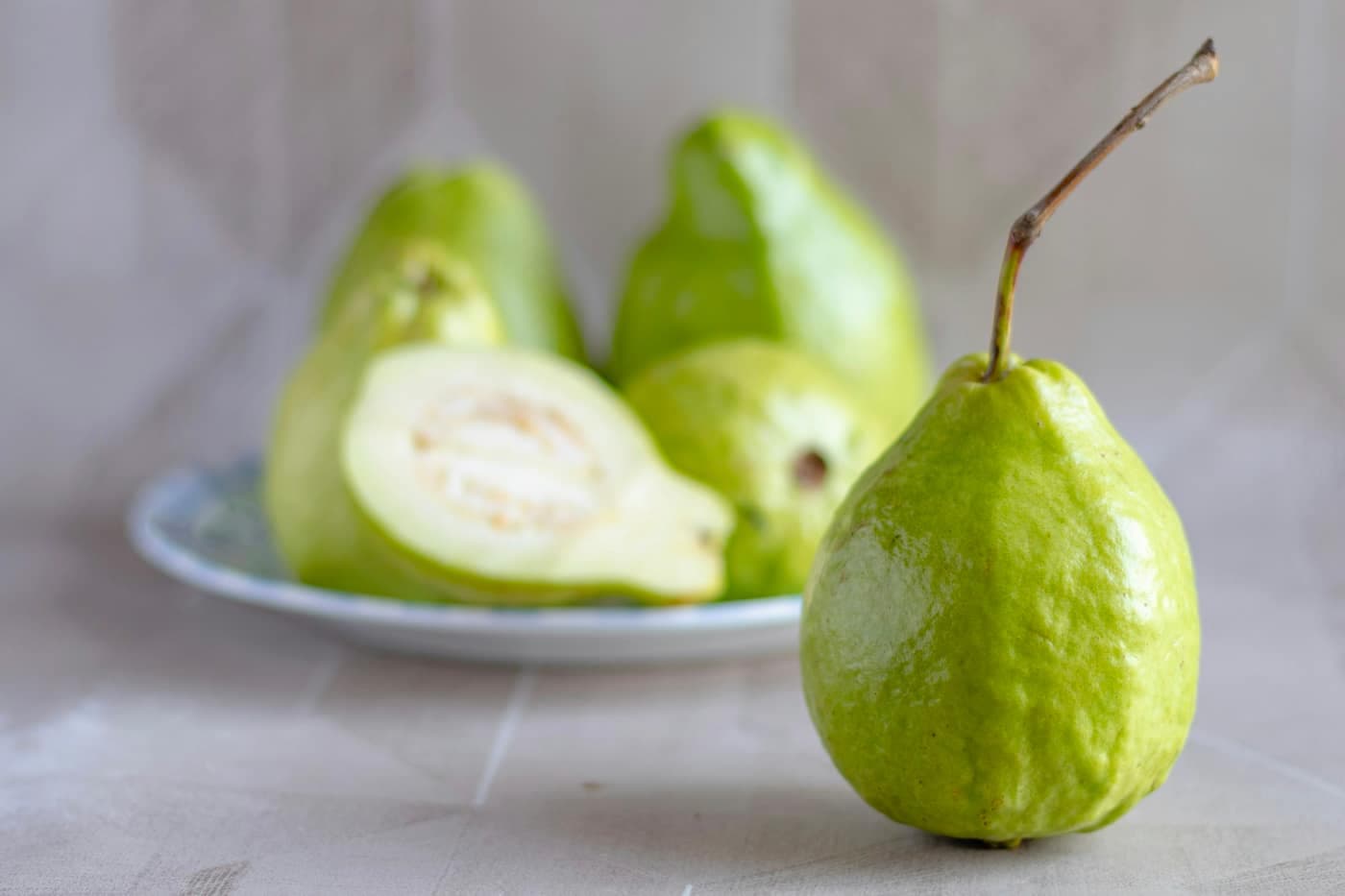A whole green guava with its stem beside a plate holding a halved guava with pale flesh