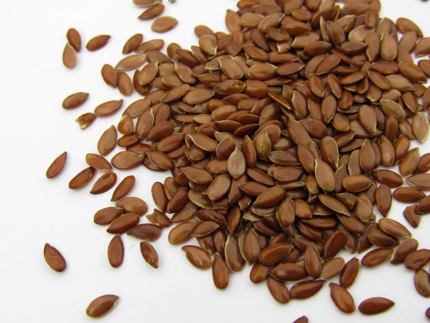 A close-up macro pile of glossy reddish-brown flax (linseed / alsi) seeds spilling across a white surface