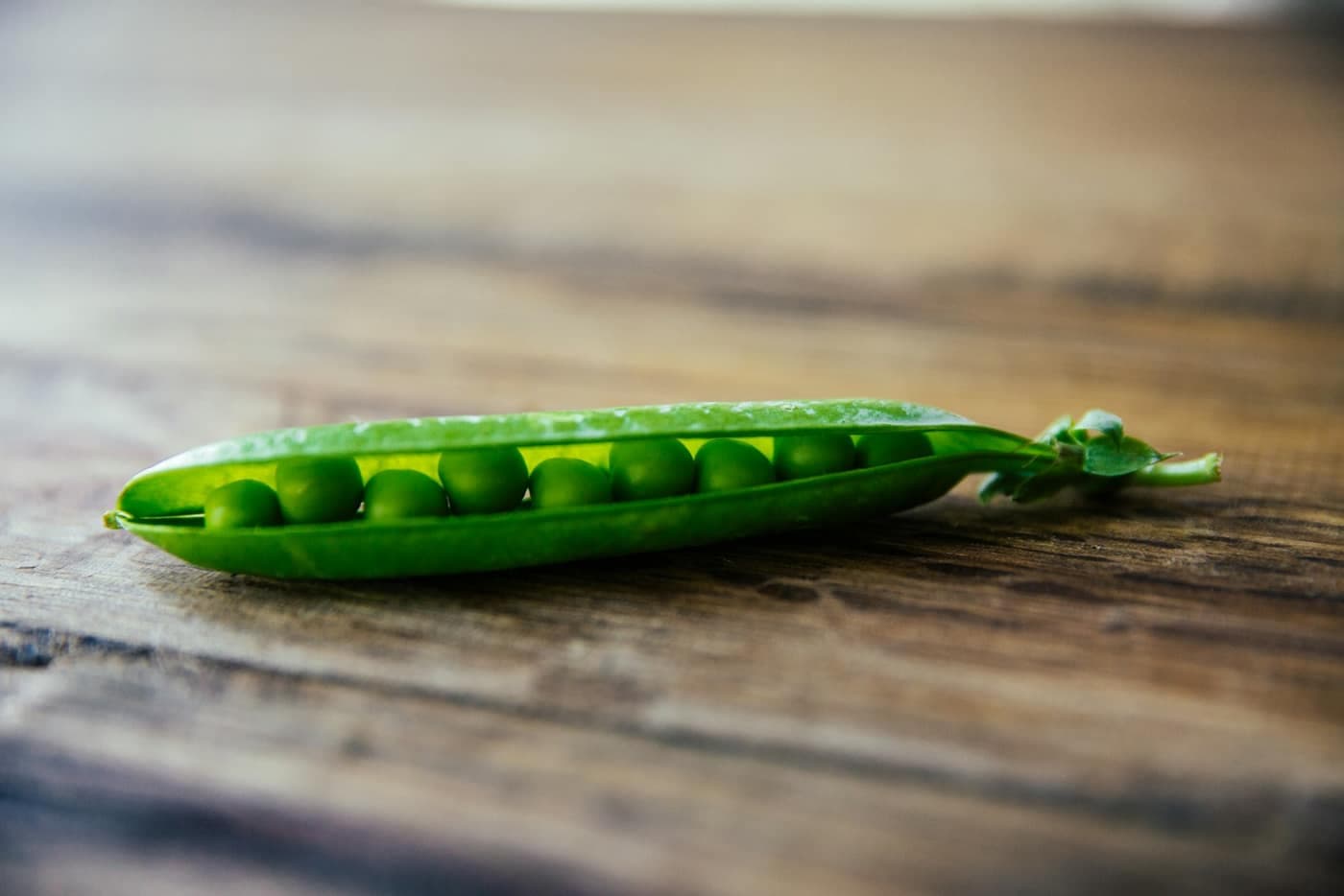 A single open green pea pod showing the peas inside, on a wooden surface