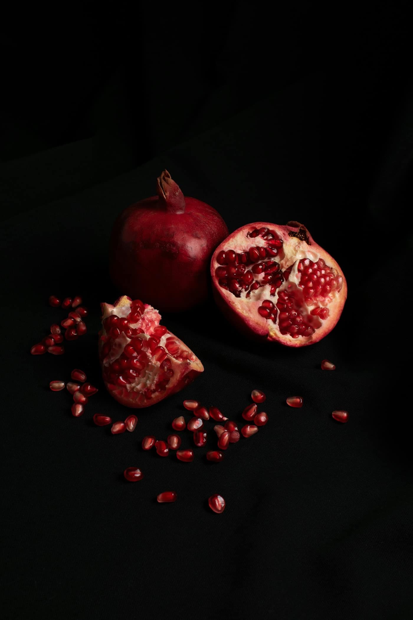A whole pomegranate beside an opened half and scattered red arils on a dark background