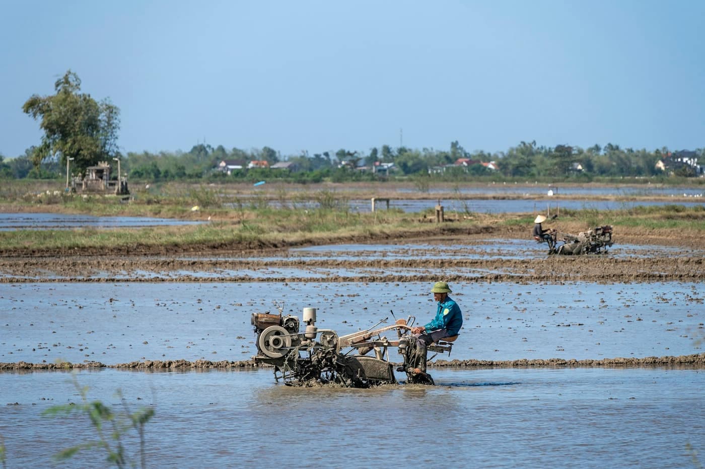 A farmer riding a walk-behind power tiller through a flooded paddy field