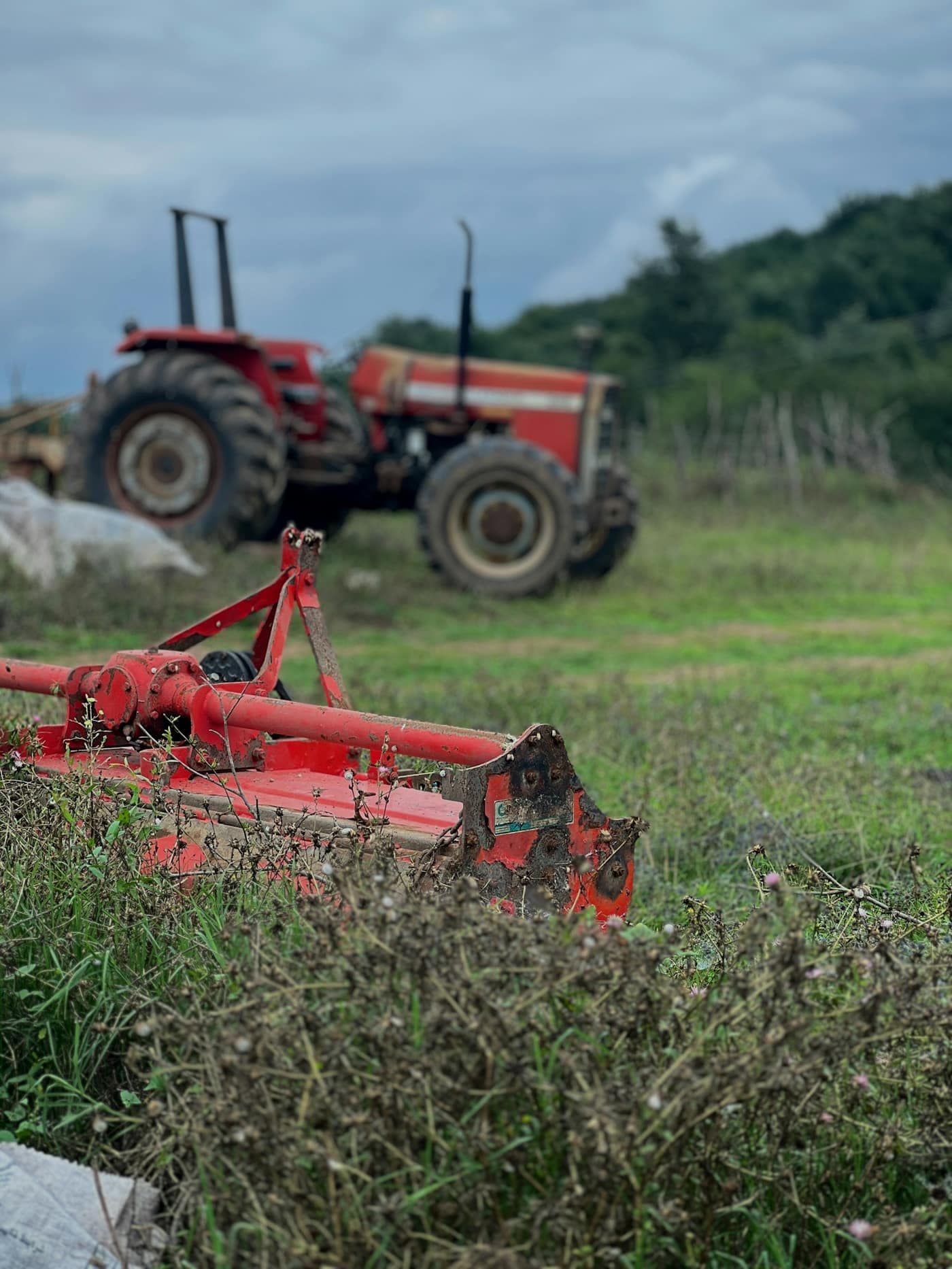 A red rotavator (rotary tiller) implement in the foreground with a tractor parked behind it in a field
