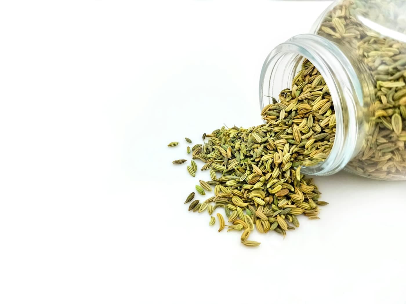 A glass jar filled with pale green, ridged fennel (saunf) seeds on a white table