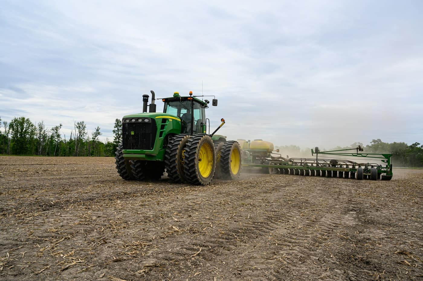 A tractor pulling a wide seed drill through a field, raising a trail of dust