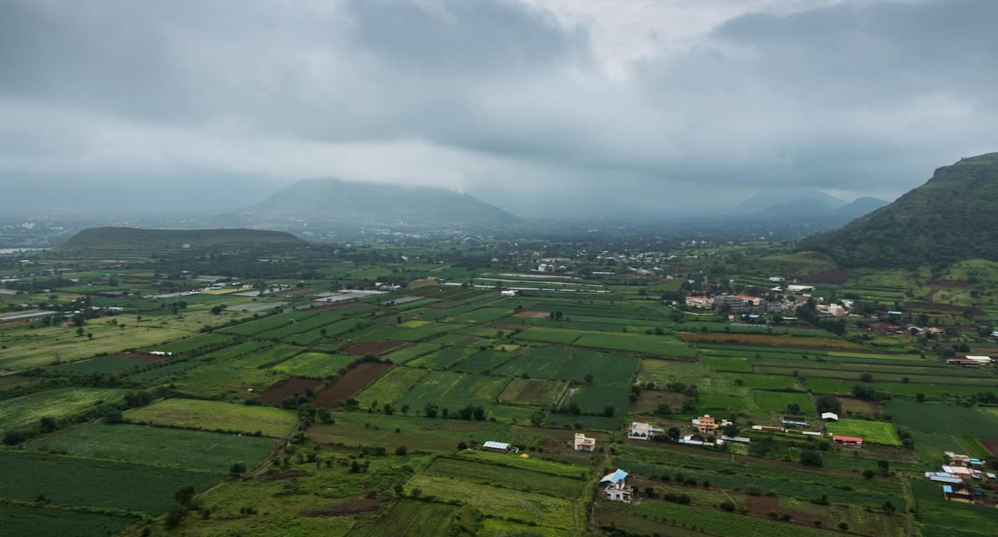 Aerial view of a patchwork of green farm fields and a small village spread across a valley between hills under a cloudy monsoon sky